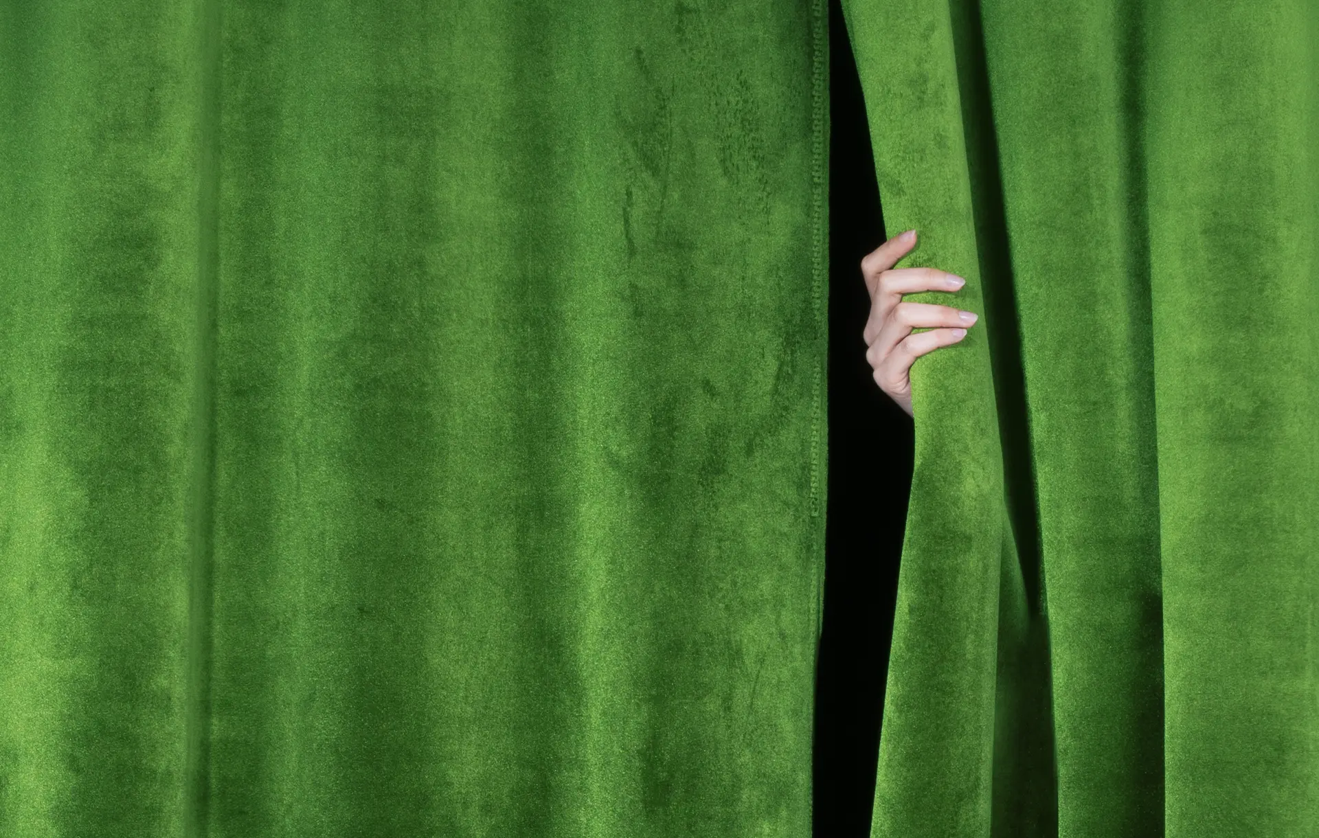 A hand lightly pulling open a green velvet stage curtain.